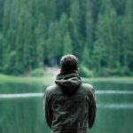 A person in a green jacket stands by a serene forest lake, capturing calming nature.